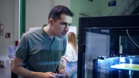 Joven-ingeniero-caminando-hacia-la-máquina-de-impresión-3D-en-laboratorio,-observando-el-proceso-de-impresión-tridimensional-y-revisando-notas-en-su-bloc-de-notas,-rastreando-la-toma