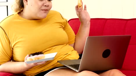 Mujer-con-sobrepeso-comiendo-rosquilla-y-usando-computadora-portátil-en-el-sofá-en-casa