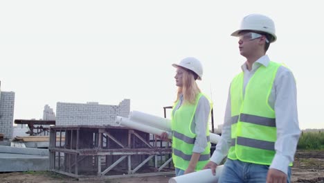 Mann-und-Frau-Betreuer-und-Assistent-in-Zeichnungen-in-weißen-Helmen-gehen-und-sprechen-auf-der-Baustelle-zeigt-die-Lage-der-Objekte