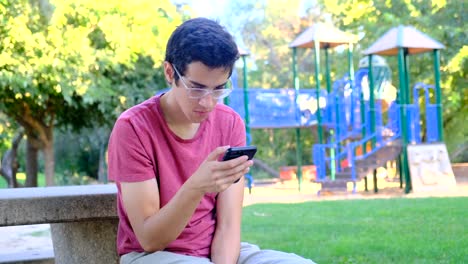 Young-man-using-his-smart-phone-sitting-in-a-park-bench