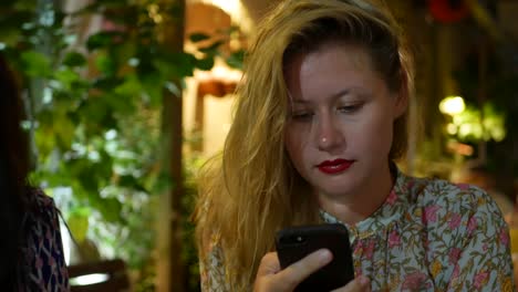 Pensive-girl-uses-a-smartphone,-writes-a-message,-chatting.-Woman-looking-for-information-in-the-phone-and-sad.-Sitting-in-restaurant-alone.-Evening-time,-lowlight.-Technology-and-entertainment-concept.-4k