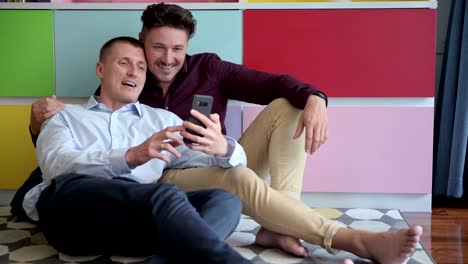 Pareja-gay-usando-teléfono-en-sillón-juntos.-Videollamada-a-amigo.