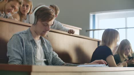 Ein-männlicher-Student-in-einem-Auditorium-der-Universität-hört-während-einer-Pause-Musik-in-weißen-Kopfhörern,-ohne-auf-andere-zu-achten.-Viele-Leute-im-Publikum-reden