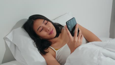 Young-Asian-woman-using-smartphone-checking-social-media-feeling-happy-smiling-while-lying-on-bed-after-wake-up-in-the-morning,-Beautiful-attractive-hispanic-girl-smiling-relax-in-bedroom-at-home.