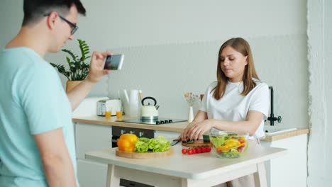 Girl-cooking-salad-talking-showing-thumbs-up-while-guy-recording-video-for-vlog