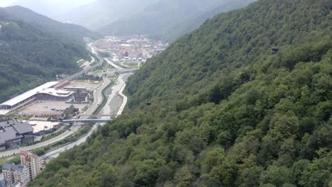 Drone-vista-cabina-del-teleférico-moviéndose-en-el-bosque-de-verano-de-montaña-entre-árboles-verdes.-Ciudad-moderna-entre-las-tierras-altas-verdes.-Arquitectura-urbana-y-río-en-valle-de-montaña