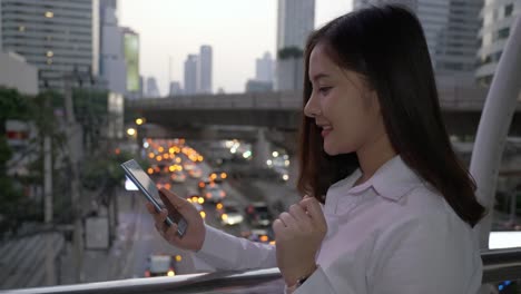 Joven-mujer-asiática-sonriente-usando-teléfono-inteligente-en-el-centro-de-la-ciudad-moderna-en-el-crepúsculo-de-Bangkok-Tailandia.-Usa-su-dedo-para-tocar-la-pantalla.-Para-usar-hologramas-Usted-diseña