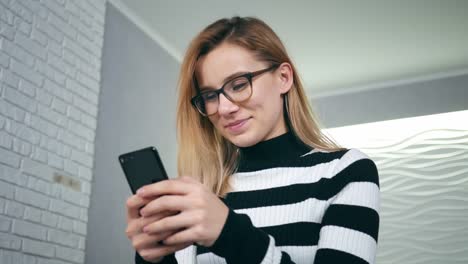 Mujer-feliz-con-teléfono-inteligente-disfrutando-de-las-redes-sociales-en-el-trabajo-tocando-la-pantalla-sonriendo-en-casa.-Mujer-feliz-con-teléfono-inteligente-está-disfrutando-de-las-redes-sociales-tocando-la-pantalla-sonriendo-sentado-en-el-sofá