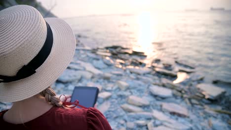 Close-up-View-from-the-back-An-attractive-young-girl-in-a-summer-red-dress-and-a-straw-hat-sits-on-a-stone-by-the-sea-at-sunset-and-watches-something-on-a-tablet.-Swipe-across-the-screen