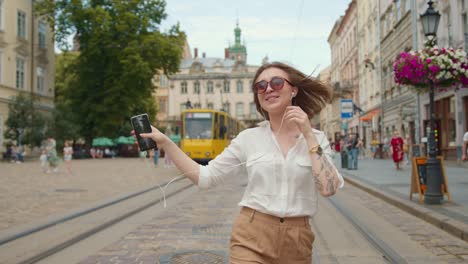 The-girl-is-going-and-listening-to-music-with-headphones.-She-is-very-happy-and-dancing.-Tram-is-riding-and-people-are-walking-in-the-background.-4K