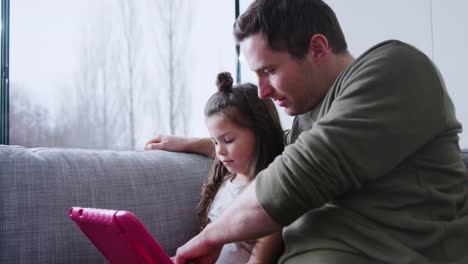 Vater-und-Tochter-sitzen-auf-Sofa-zu-Hause-spielen-zusammen-auf-digitalen-Tablet-in-rosa-Fall-zu-Hause