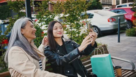 Asian-mature-woman-and-her-daughter-are-smiling-and-showing-victory-sign,-taking-selfie-by-smartphone-while-sitting-outdoors-on-a-bench