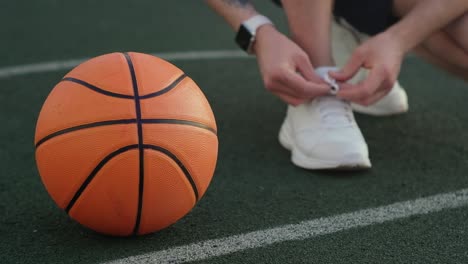 Unbekannter-Sportler-bindet-Schnürsenkel-an-Turnschuhen,-die-in-der-Nähe-eines-Basketballballs-posieren