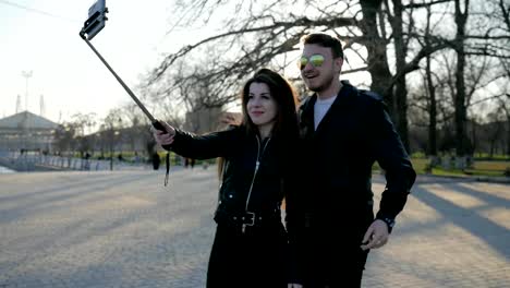 couple-takes-photo-on-mobile-gadget-in-selfie-stick,-phone-on-self-stick,-stylish-guy-girl-walk-in-park