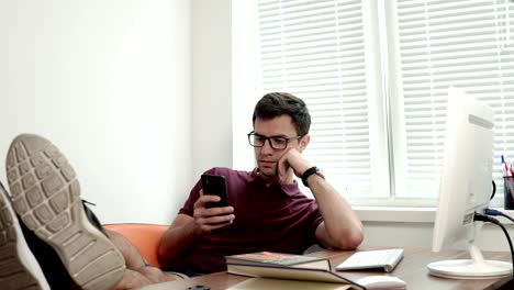 Young-worker-in-glasses-relaxing-with-his-legs-on-the-table-modern-office-using-phone.-Bored-manager-surfing-in-internet.-Shot-4k