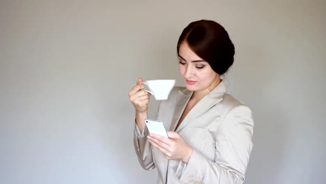 Beautiful-young-businesswoman-looks-at-the-phone-screen-and-plays-downloaded-application-on-a-smart-phone.-Elegant-woman-communicate-in-social-networks-and-drinks-coffee