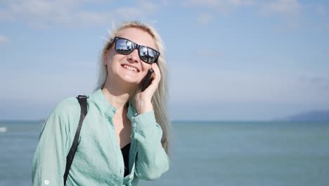 Beautiful-slim-woman-with-long-blonde-hair-and-green-shirt-standing-and-talking-on-phone-over-background-sea