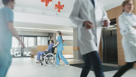 First-Floor-of-the-Busy-Hospital,-Doctors,-Nurses-and-Personnel-Busy-Working,-Receptionist-Talks-with-a-Patient,-Assistant-Moves-Elderly-Man-in-the-Wheelchair.-New-Modern-Medical-Hospital-with-Professional-Staff.