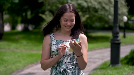 Smiling-asian-girl-goes-along-the-paved-track-in-the-park.-Chatting-in-social-networks-on-her-yellow-smartphone.