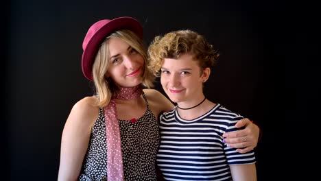 Two-young-beautiful-blonde-woman-hugging-and-smiling-at-camera,-isolated-on-black-background