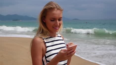 Young-beautiful-slim-woman-with-long-blonde-hair-in-black-and-white-dress-standing-on-the-coast-and-using-smartphone-over-background-at-storm-on-the-sea..-Girl-on-the-beach-touching-screen-and-smile.