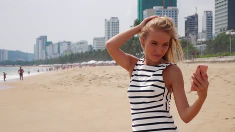 Young-beautiful-slim-woman-with-long-blonde-hair-in-black-and-white-dress-making-selfie-on-mobile-phone-near-the-sea-on-background-of-city