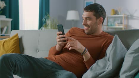 Handsome-Happy-Man-at-Home,-Sits-Down-on-a-Sofa-while-Using-Smartphone,-Does-Various-Touching-and-Swiping-Gestures.-Man-Relaxing-with-Mobile-Phone-in-His-Cozy-Living-Room.