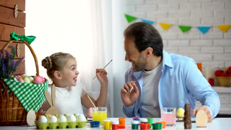 Happy-daughter-and-father-having-fun-painting-each-other-with-brush,-Easter