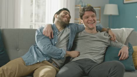 Sweet-Male-Gay-Couple-Full-Around-on-a-Sofa-at-Home.-Boyfriend-Runs-and-Jumps-into-Hands-of-His-Partner.-They-Hug.-They-are-Happy-and-Laughing.-They-are-Casually-Dressed-and-Room-Has-Modern-Interior.