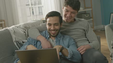 Dulce-pareja-gay-hombre-pasa-tiempo-en-casa.-Son-de-mentira-abajo-en-un-sofá-y-usar-la-Laptop.-Navegan-en-línea.-Mano-de-socio-es-alrededor-de-su-amante.-Sonreír-y-reír.-Habitación-tiene-un-Interior-moderno.