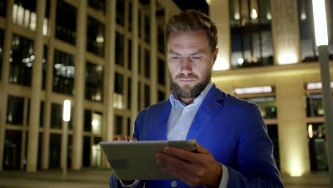 Medium-shot-of-serious-businessman-using-tablet-computer-in-illuminated-might-street