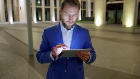 Schräg-nach-unten-mittlere-Aufnahme-von-ernsthaften-Geschäftsmann-Surfen-Internet-auf-dem-Tablet-PC-während-des-Gehens-in-Nacht-Stadtstraße