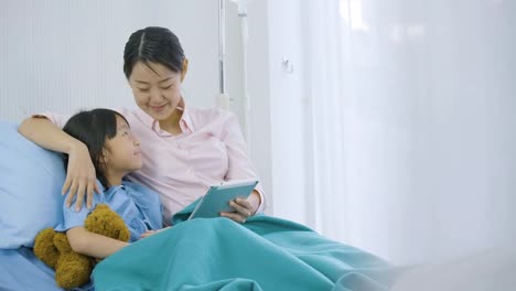 Cute-little-girl-lying-on-bed-with-her-mother-in-hospital,-watching-funny-cartoons,-movies-on-digital-tablet.-illness-and-treatment.