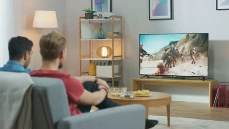 Im-Wohnzimmer:-Zwei-Freunde-entspannen-sich-auf-einem-Couch-Watching-War-Movie-auf-einem-Fernseher-Moderne-militärische-Kriegshilfe-mit-Schießen-und-Explosionen-auf-einem-Fernseher.