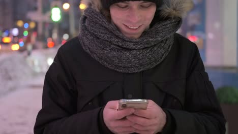 Hombre-caminando-en-la-calle-en-ropa-de-abrigo-y-mensajería-en-el-teléfono-móvil-por-la-noche