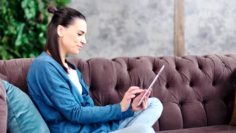 Side-view-laughing-teenage-young-girl-using-tablet-pc-sitting-on-couch-at-cosiness-living-room