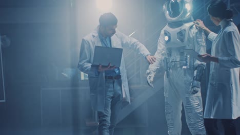 Diverse-Teams-von-Luft-und-Raumfahrt-Wissenschaftlern-und-Ingenieuren,-die-weiße-Mäntel-tragen,-haben-Diskussionen,-Benutzung-von-Computern,-Design-New-Space-Suit,-die-für-Galaxy-Exploration-und-Reisen-angepasst-sind.-Astronaut-bauen-Anzug