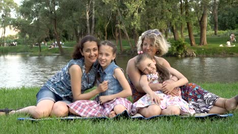 Same-sex-family-with-children-playing-and-resting-in-nature.-Two-mothers-with-young-daughters-hugging-and-having-fun,-sitting-on-the-lawn-in-the-Park-at-sunset-on-a-summer-day-on-the-river-Bank.-4K.