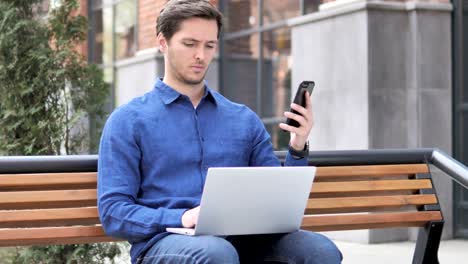 Hombre-joven-usando-smartphone-y-laptop,-sentado-en-el-Banco