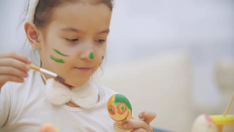 Bonita-y-linda-chica-se-está-divirtiendo-pintando-un-huevo-de-Pascua.-Adorable-chica-está-coloreando-huevo-de-Ester.-Chica-con-manchas-de-belleza-en-su-cara-y-está-sonriendo-suavemente-con-bigote-de-gato,-sentado-en-la-mesa-de-madera-con-decoraciones-de