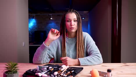 Nahaufnahme-von-jungen,-attraktiven-Make-up-Künstlerin-mit-Dreadlocks,-die-live-und-Werbekosmetik-mit-dem-Neonhintergrund-im-Haus-streamen