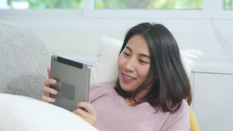 Asiatische-Frau-musik-hören-und-Tablet,-Frauen-mit-Entspannung-Zeit-liegen-auf-dem-heimischen-Sofa-im-Wohnzimmer-zu-Hause.-Fröhliche-weibliche-Hörmusik-mit-Kopfhörer-Konzept.