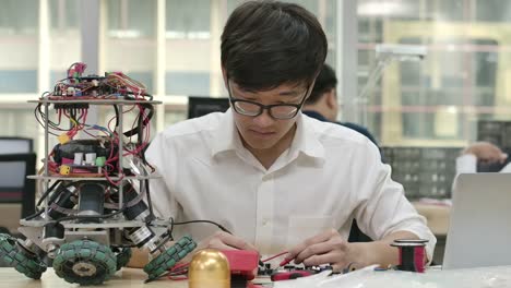 Young-asian-electronics-development-engineers-works-with-robot,-measuring-the-signal-in-the-electrical-circuits-of-robotics-prototype-in-workshop.-People-with-technology-or-innovation-concept.