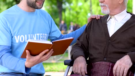 Male-volunteer-reading-book-to-lonely-disabled-pensioner-in-park,-social-support