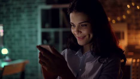 Cheerful-young-woman-using-smartphone-at-night-in-dark-office-smiling