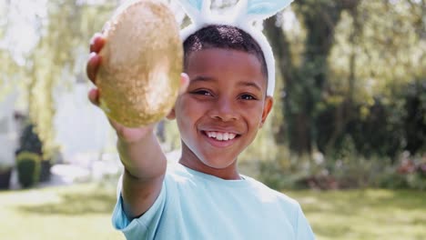 Retrato-de-niño-usando-orejas-de-conejo-en-la-caza-de-huevos-de-Pascua-al-aire-libre-en-casa-sosteniendo-huevo-de-chocolate-a-la-cámara-y-sonriendo---disparado-en-cámara-lenta