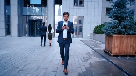 Young-Business-Man-Uses-Smartphone-While-Walking-on-the-Big-City-Business-District-Street.-Classically-Dressed.-Big-Office-Building-in-the-Background.-Looking-Successful,-Confident.