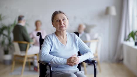 Tracking-shot-of-disabled-senior-woman-in-wheelchair-looking-away-thoughtfully,-then-turning-head-and-looking-at-camera-with-sadness-in-nursing-home,-other-aged-patients-in-background