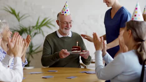 Älterer-Mann-spielen-Karten-mit-alten-Freunden-in-Party-Hüte,-während-Betreuer-bringt-ihm-Geburtstagstorte.-Glücklicher-Senior-Mann-immer-Wünsche-und-Umarmungen-und-Blasen-Kerze-aus-im-Pflegeheim