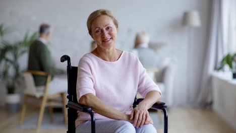 Tracking-shot-of-disabled-senior-woman-in-wheelchair-looking-away-thoughtfully,-then-turning-head,-smiling-and-laughing-at-camera-happily-in-nursing-home,-other-aged-patients-in-background